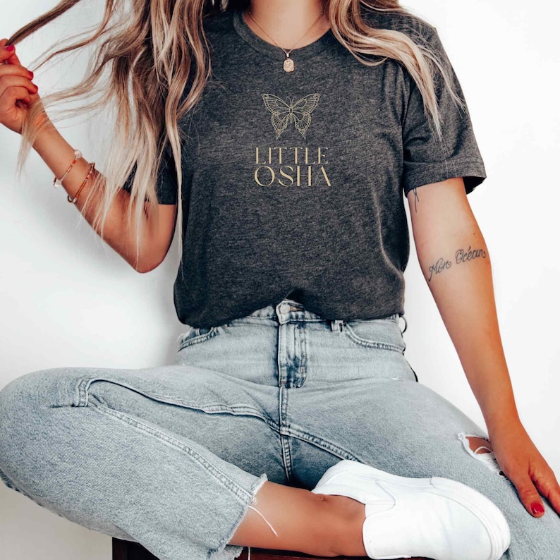 May include: A dark gray t-shirt with a gold butterfly graphic and the text "LITTLE OSHA" printed on the front.