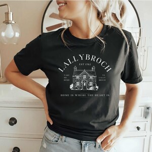 May include: Black t-shirt with a white graphic design of a house with the text "Lallybroch Est 1702" above the house and "Take me home" and "To Lallybroch" on either side of the house. The text "Home is where the heart is" is below the house.