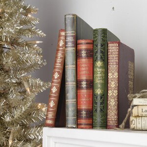 May include: A collection of vintage hardcover books with ornate designs and gold lettering. The books are arranged on a white shelf with a stack of old newspapers tied with twine on the right.