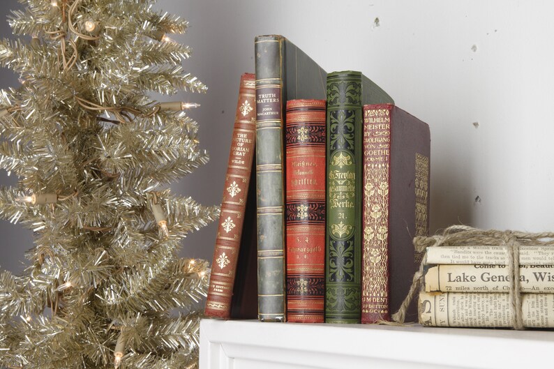 May include: A collection of vintage hardcover books with ornate designs and gold lettering. The books are arranged on a white shelf with a stack of old newspapers tied with twine on the right.
