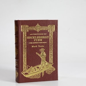 May include: A red hardcover book with gold lettering titled "Adventures of Huckleberry Finn (Tom Sawyer's Companion)" by Mark Twain. The cover features an illustration of a man in a boat.