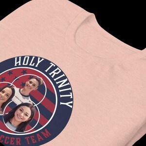 以下が含まれることがあります： ピンクのTシャツに、ネイビーブルーとレッドの円形のグラフィックがプリントされています。グラフィックには「HOLY TRINITY SOCCER TEAM」というテキストと、女性の顔が3つ描かれています。