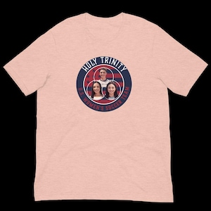 以下が含まれることがあります： ピンクのTシャツに、ネイビーブルーと白の文字で「Holy Trinity U.S. Women's Soccer Team」と書かれた円形のデザインが施されています。デザインには、赤、白、青のアメリカ国旗と、女性の顔が3つ描かれています。