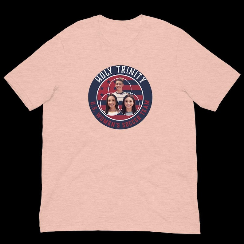 以下が含まれることがあります： ピンクのTシャツに、ネイビーブルーと白の文字で「Holy Trinity U.S. Women's Soccer Team」と書かれた円形のデザインが施されています。デザインには、赤、白、青のアメリカ国旗と、女性の顔が3つ描かれています。