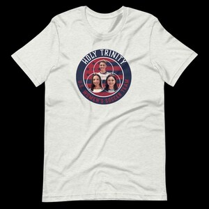以下が含まれることがあります： 3人の女性の顔が描かれた青い円形のグラフィックと「HOLY TRINITY U.S. WOMEN'S SOCCER TEAM」の文字が書かれた白いTシャツ。