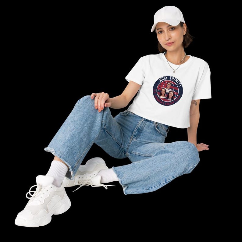 May include: White crop top with a circular graphic that reads "Holy Trinity" and features three faces. The model is wearing light blue jeans, white sneakers, and a white baseball cap. The background is black.