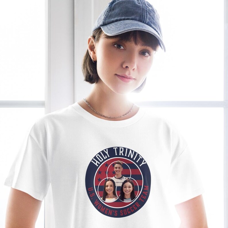 May include: White t-shirt featuring a circular graphic with the text "HOLY TRINITY" and "U.S. WOMEN'S SOCCER TEAM". The graphic includes images of three people within a red, white, and blue design. The person in the image is wearing a blue denim baseball cap.