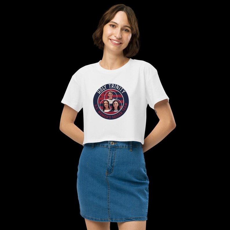 May include: White crop top with a circular graphic featuring the text "HOLY TRINITY" and "WOMEN'S SOCCER TEAM". The graphic includes images of three women. The top is paired with a denim skirt.