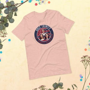 以下が含まれることがあります： 3人の女性の顔が描かれた円形のグラフィックと「Holy Trinity US Women's Soccer Team」というテキストがプリントされたピンクのTシャツ。
