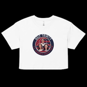 May include: White crop top featuring a circular graphic with the text "HOLY TRINITY U.S. WOMEN'S SOCCER TEAM." The graphic includes images of three women and a red, white, and blue color scheme. The shirt has short sleeves and a crew neck.