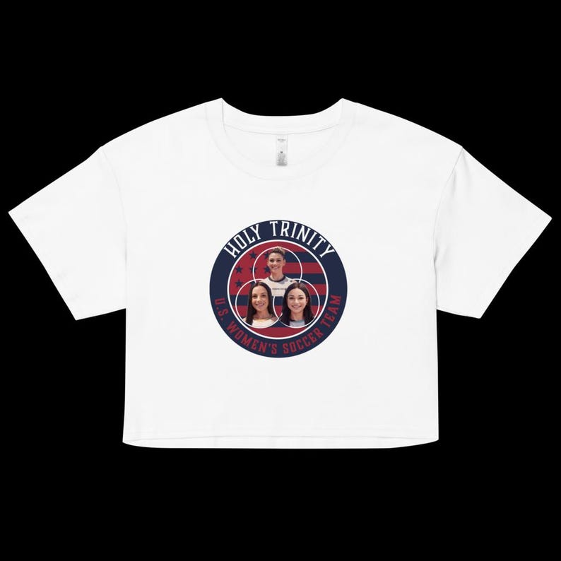 May include: White crop top featuring a circular graphic with the text "HOLY TRINITY U.S. WOMEN'S SOCCER TEAM." The graphic includes images of three women and a red, white, and blue color scheme. The shirt has short sleeves and a crew neck.