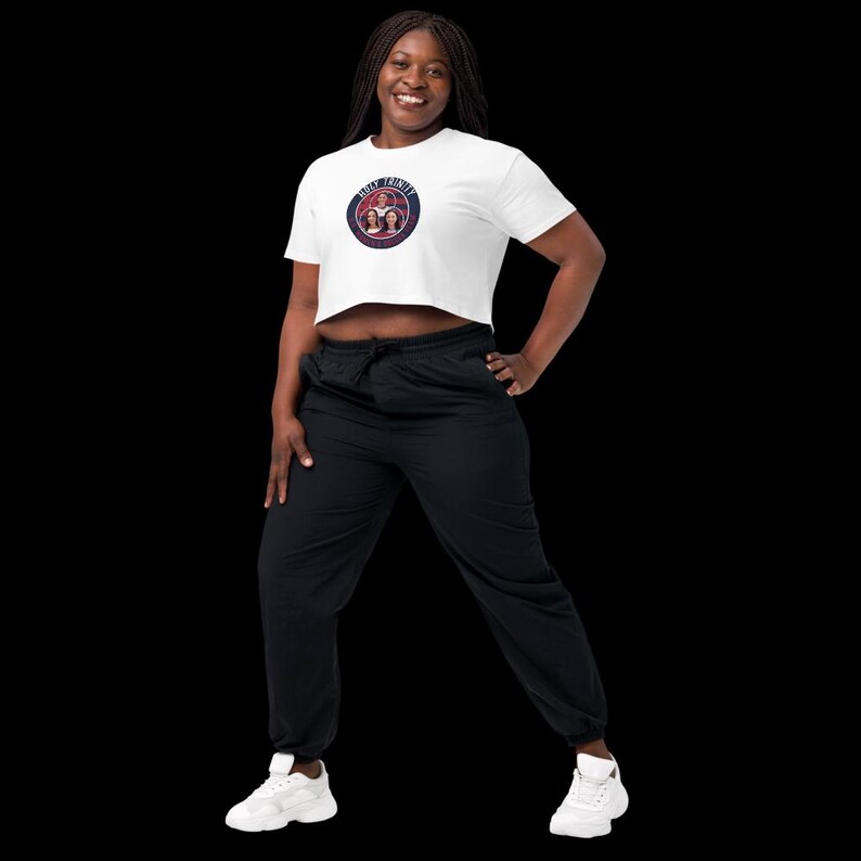 May include: A white crop top with a circular graphic that reads "Holy Trinity" in a navy blue font. The top is paired with black sweatpants and white sneakers. The person is smiling and posing against a black background.