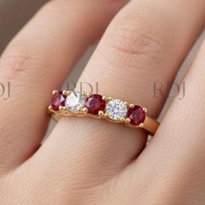 May include: A gold band ring featuring a row of alternating red gemstones and clear gemstones. The ring is set on a hand, showcasing the design and sparkle of the jewelry. The gemstones are round cut.