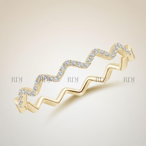 May include: Two gold-colored rings with a zigzag design. One ring is fully gold, while the other features a zigzag pattern of small, clear gemstones. The rings are set against a light beige background.