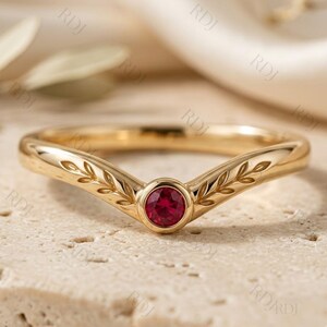 May include: A gold V-shaped ring featuring a central round-cut red ruby gemstone. The band is decorated with an engraved leaf pattern extending from the centre stone along both sides of the chevron-shaped shank, creating a polished finish.