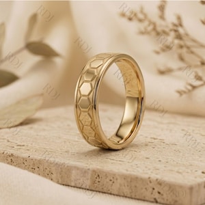 May include: A gold-toned ring featuring a repeating geometric honeycomb pattern engraved around the entire outer band. The ring has a polished finish with a smooth interior surface and a consistent width, showcasing a detailed hexagonal texture.