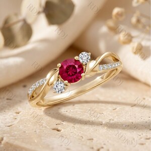 Peut inclure: Une bague en or ornée d'une pierre précieuse rouge vif ronde au centre, entourée de deux petites pierres blanches rondes. L'anneau présente une finition dorée polie avec des motifs en forme de feuilles et des petits pavés de pierres claires.