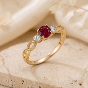 Puede incluir: Un anillo de oro con una piedra central de rubí rojo intenso de corte redondo, flanqueada por dos diamantes blancos redondos más pequeños. La banda presenta un diseño calado trenzado con delicadas piedras en pavé.