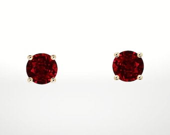 May include: A pair of round, deep red gemstone stud earrings. Each earring has a vibrant red stone set in a gold-coloured, four-prong setting. The earrings are shown against a plain white background.