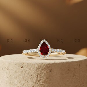 Op de afbeelding: Een gouden ring met een peervormige, dieprode edelsteen, omringd door een halo van kleine, heldere stenen. De band is ook versierd met kleine, heldere stenen. De ring wordt gepresenteerd tegen een neutrale achtergrond.