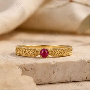 May include: A gold ring featuring a central round-cut red gemstone. The band is crafted with a detailed leaf or wheat pattern along the shank, bordered by a beaded edge, creating a textured appearance across the entire circumference of the band.