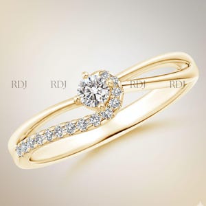 Op de afbeelding: Een goudkleurige ring met een centrale, ronde diamant en kleinere diamanten langs een gebogen band. De ring heeft een uniek, open ontwerp met een wervelende accent rond de hoofdsteen. Zachte, neutrale achtergrond.