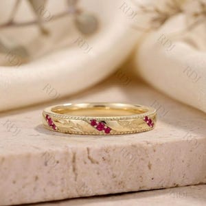 May include: A gold band ring featuring a series of small, round, vibrant red rubies set in a diagonal pattern. The metal surface is decorated with engraved leaf motifs and a delicate beaded border along the top and bottom edges of the band.