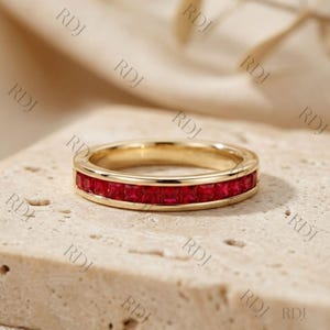 May include: A gold band ring with a row of square-cut, vibrant red gemstones. The ring is set on a neutral-toned surface, showcasing the ring's design and color. The ring is a piece of jewelry.