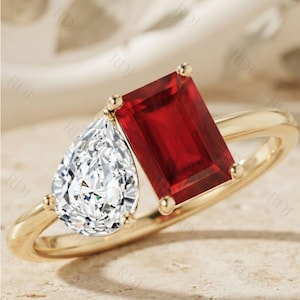 Pode incluir: Um anel de ouro com uma pedra preciosa vermelha retangular de corte esmeralda e uma pedra preciosa branca transparente em forma de pera lado a lado. A banda é de ouro amarelo polido e as pedras estão presas com pequenas garras de ouro.