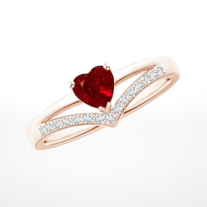 Puede incluir: Anillo de oro rosa con una piedra preciosa roja oscura en forma de corazón. El anillo tiene un diseño de caña dividida, con una fila de pequeñas piedras preciosas claras engastadas a lo largo de la banda exterior. La piedra en forma de corazón es el punto focal.
