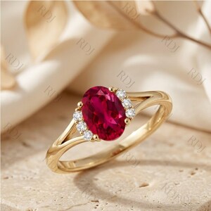 May include: A gold ring featuring a large oval-cut deep red gemstone at the centre, flanked by three small round white accent stones on each side. The band has a split-shank design, and the metal has a polished, reflective yellow gold finish.