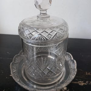Puede incluir: Una mantequera de cristal transparente con tapa y base decorativa. La tapa tiene un pomo facetado y patrones tallados en forma de diamante. El plato se apoya en una base redonda con bordes festoneados. El cristal tiene un aspecto vintage y elegante.