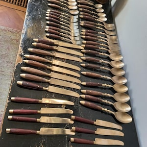 Puede incluir: Una colección de cubiertos antiguos, que incluye cuchillos, tenedores y cucharas, dispuestos sobre una superficie oscura. Los utensilios presentan hojas y púas de latón, con mangos de madera oscura y detalles de latón. Las cucharas tienen un diseño retorcido.