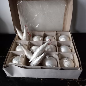 May include: A cardboard box filled with vintage Christmas ornaments. The box contains white, pearlescent ornaments in various shapes, including teardrops and spheres. Each ornament has a silver cap and a red string for hanging. The box is divided into compartments.