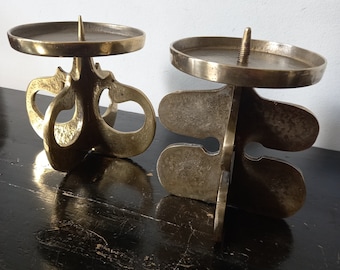 May include: Two gold metal candle holders with abstract, organic shapes. The holders have a round top and a base with multiple curves and loops.