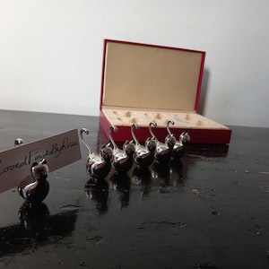 May include: A set of silver-coloured swan-shaped place card holders, with a cat-shaped holder in front holding a card that reads "PreLoved Finds by Roza". A red box with a beige interior is in the background.