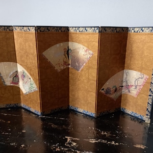 Peut inclure: Un paravent pliant à six panneaux avec un fond doré texturé et une bordure florale bleu foncé. Trois illustrations en forme d'éventail sont disposées sur les panneaux, représentant des personnages dans des paysages avec des arbres.