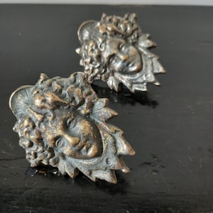 Può includere: Due maniglie antiche per armadi in bronzo. Ogni maniglia presenta un volto dettagliato con capelli ricci e un design a foglie attorno al volto. Le maniglie hanno un aspetto vintage e invecchiato, con un colore bronzo scuro.