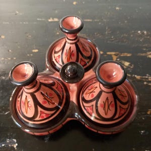 May include: A ceramic triple condiment server featuring three connected bowls with conical lids. The piece is glazed in a dusty pink hue with black painted line details and small orange and red accents on the lids and base rims.