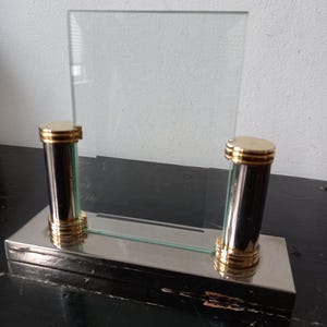 May include: A silver and gold picture frame with a glass pane. The frame has two columns with gold accents and a rectangular base.