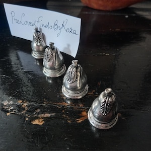 May include: Four silver-colored pear-shaped place card holders with a leaf design on a dark, distressed wooden surface. The holders have a slot for cards. A handwritten note reads "Preloved Finds By Roza".