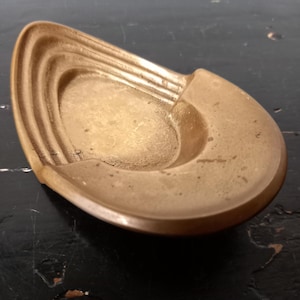 May include: A vintage, teardrop-shaped brass ashtray. The ashtray features a shallow, oval basin with a series of curved ridges on one side. The brass has a warm, aged patina, suggesting its vintage origin. The ashtray is designed to sit flat on a surface.