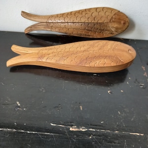May include: Two wooden fish-shaped boxes. The boxes are carved from light brown wood, with detailed scale patterns and fin designs. One box is open, revealing the interior. The boxes are displayed on a dark surface.