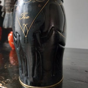 May include: Black tea tin with gold lettering and an embossed elephant design. The tin reads "Williamson &" on the lid and "Tea" on the front. The tea container has a cylindrical shape with a gold rim.