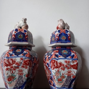 May include: A pair of hexagonal ceramic ginger jars featuring intricate red, blue, and white floral and architectural patterns. Each jar is topped with a removable lid featuring a sculpted animal finial, showcasing detailed hand-painted motifs.