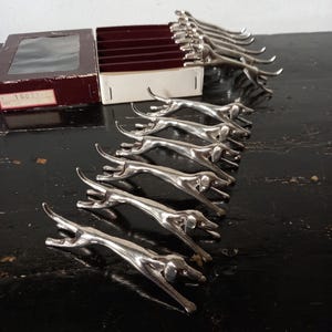 May include: A set of silver-colored dog-shaped place card holders. The dogs are in a running pose, with their legs extended. The place card holders are arranged in a row, with a burgundy box in the background.