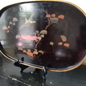 Peut inclure: Un plateau ovale en laque noire avec un bord doré et un motif peint représentant un oiseau en vol parmi des branches stylisées aux feuilles roses et dorées. Le plateau est présenté sur un support en plastique noir, révélant sa finition brillante.