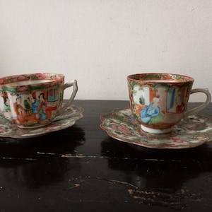 May include: Two antique porcelain teacups and saucers. The cups and saucers are decorated with colourful scenes of people and floral patterns. The rims are trimmed with gold. The saucers have a scalloped edge.