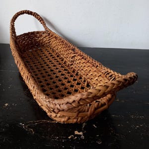 Puede incluir: Una cesta rectangular tejida marrón con asa. La cesta tiene una forma larga y estrecha con un tejido en forma de diamante. El asa es arqueada y está unida a la parte superior. Hecha de materiales naturales.