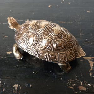 May include: A bronze-colored turtle figurine with a detailed shell pattern. The turtle has a textured shell and is posed on a dark surface. The figurine is a decorative item.